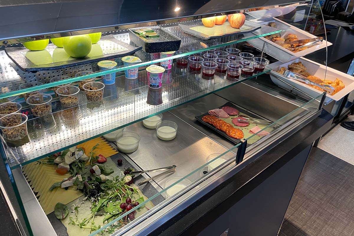 Eine gekühlte Vitrine mit Äpfeln, Bechern mit Joghurt und Müsli, Gläsern mit Saft, einer Platte Aufschnitt, Croissants und Salat