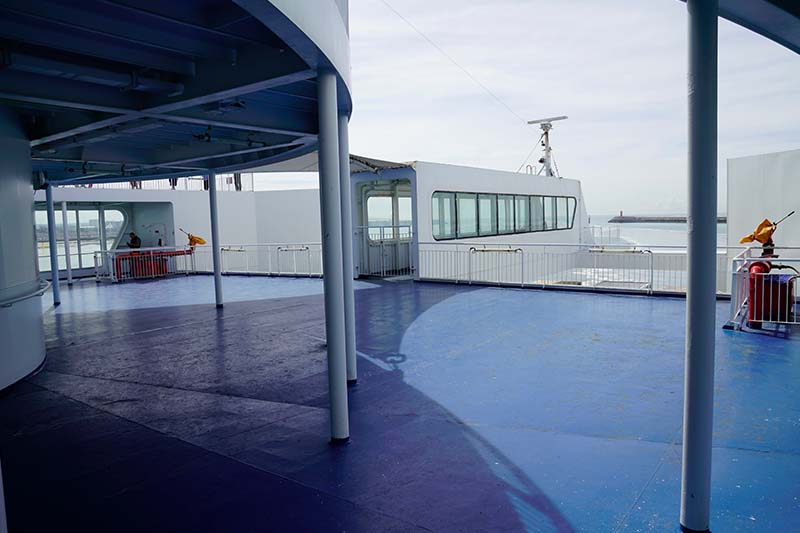 Leeres Außendeck mit blauem Boden, weißen Pfosten und Geländern und Blick auf die Hafeneinfahrt, ohne eine einzige Bank oder einen Stuhl