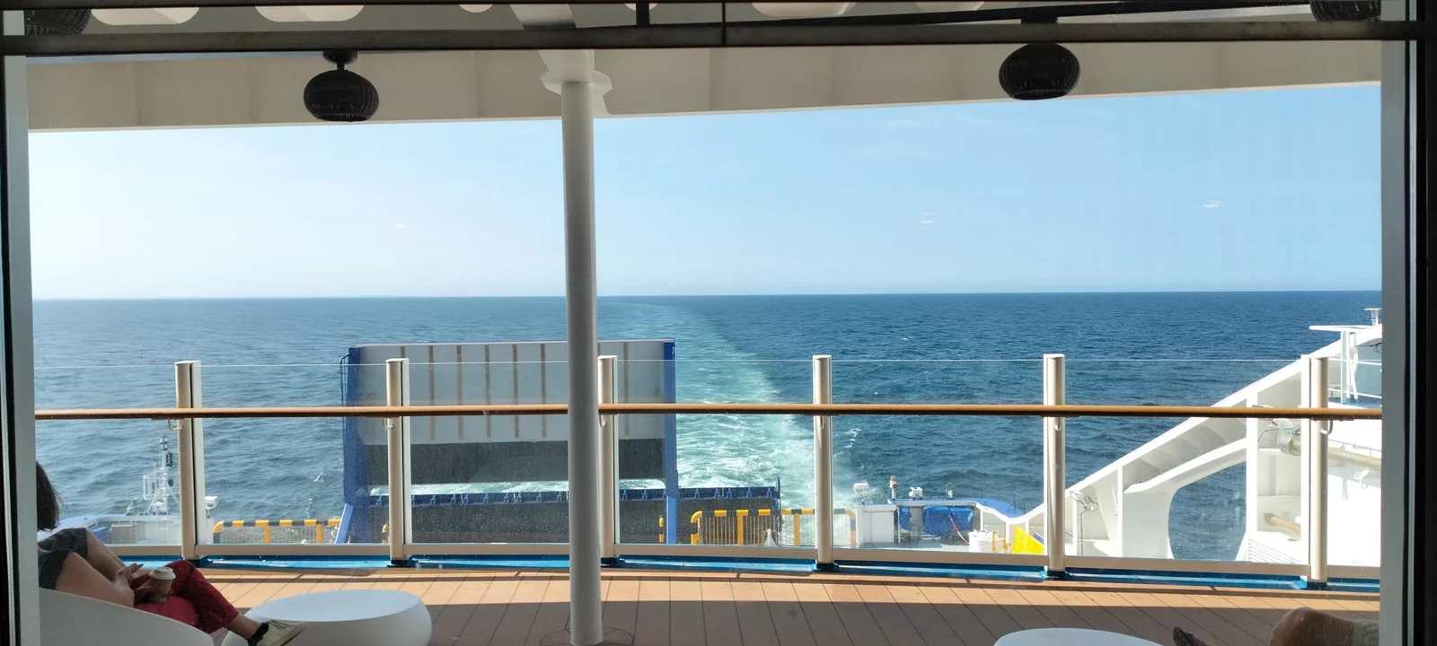 Blick vom überdachten Achterdeck der Fähre über die Reling auf das Kielwasser und das Meer, links ein Passagier in einem weißen Sessel
