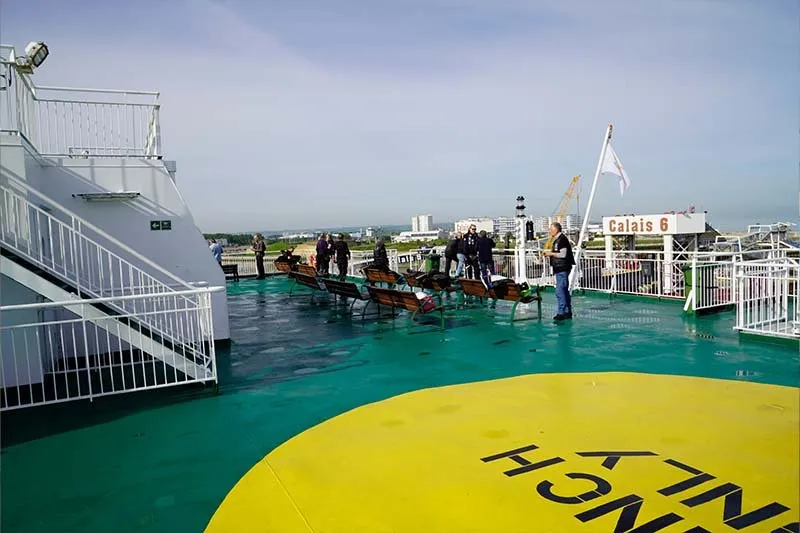 Außendeck mit Holzbänken und Menschen an der Reling, einem gelben Kreis auf dem Deckboden und am Kai dem Schild Calais 6