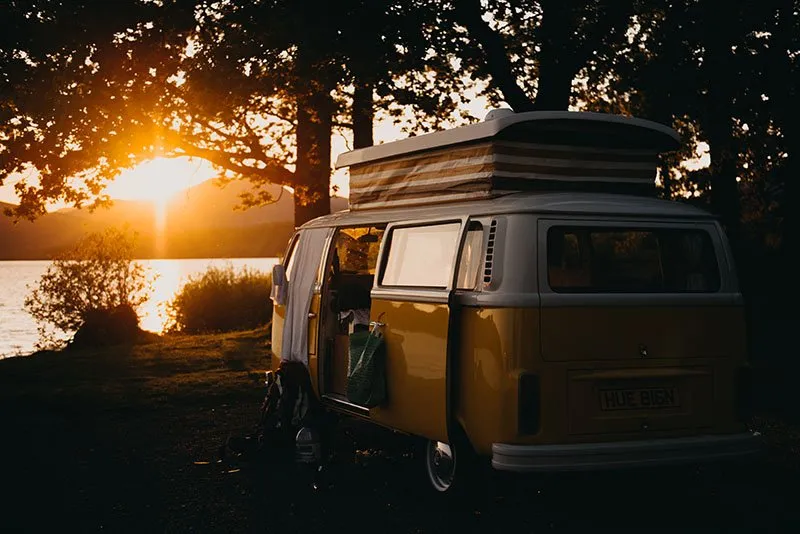 Ein alter Volkswagen-Campingbus mit Aufstelldach und britischem Kennzeichen, unter Bäumen am Ufer eines Sees bei Sonnenuntergang geparkt