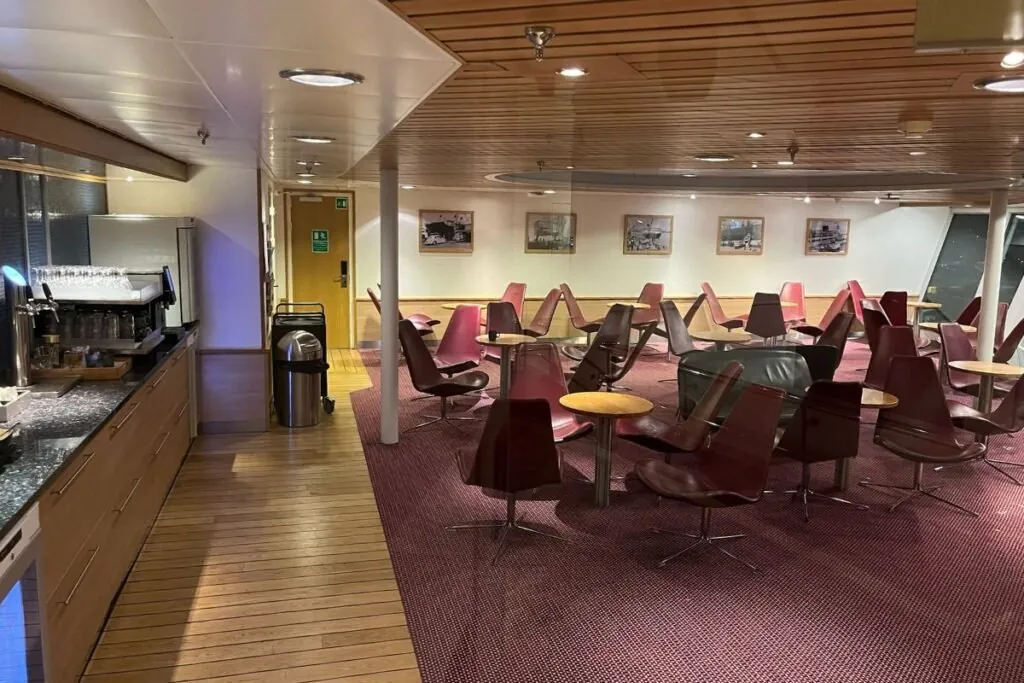 Die Commodore Lounge an Bord: rote und braune Drehsessel um kleine Tische, ein Holzboden und eine Bar mit Kaffeemaschine