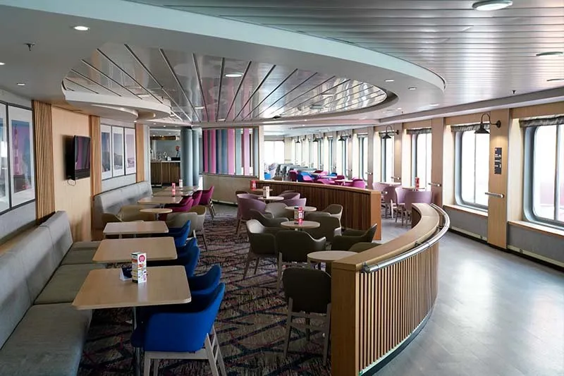Salon an Bord von DFDS mit blauen und rosa Sesseln, Holztischen und einer Bank an der Wand unter den Fenstern