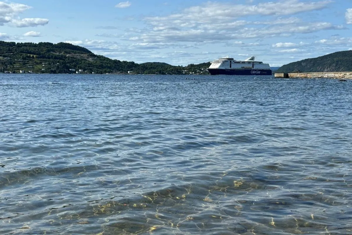 Eine Kreuzfahrtfähre von Color Line fährt durch den Oslofjord, vom Ufer aus gesehen, mit klarem flachem Wasser und bewaldeten Hügeln auf beiden Seiten