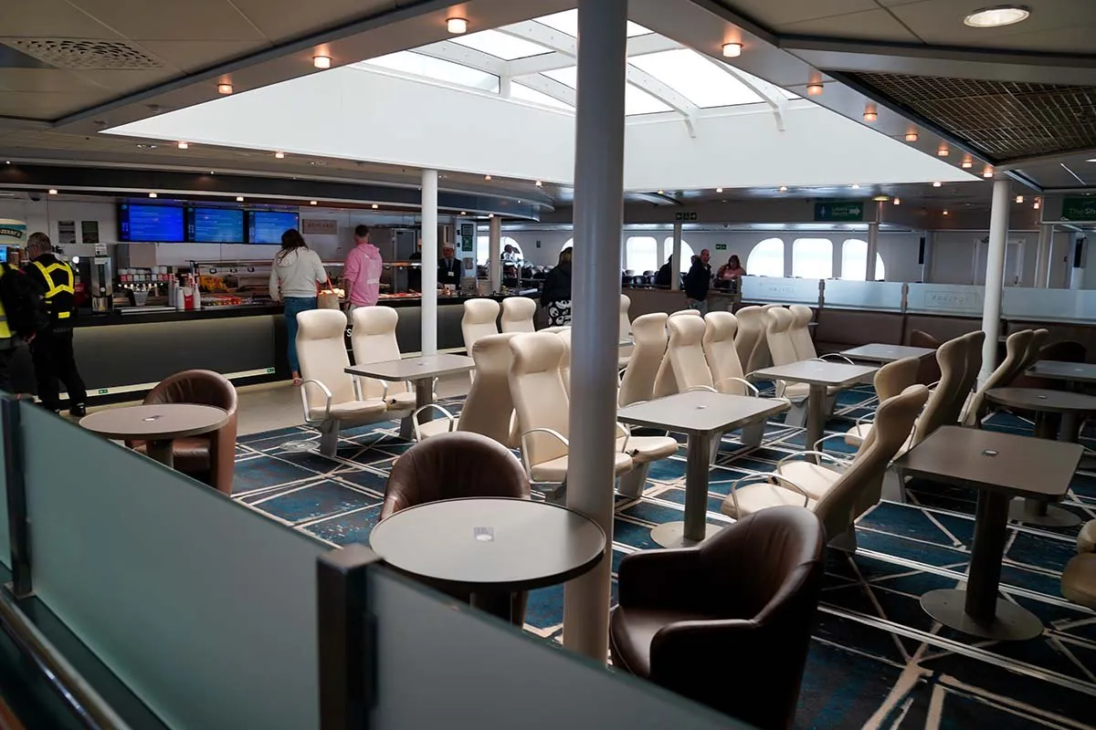 Passagierdeck mit Reihen cremefarbener Sessel, einzelnen Sesseln an runden Tischen und hinten einer Bar, an der Leute warten