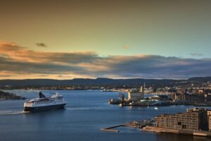 Eine Fähre in den früheren blau-weißen DFDS-Farben verlässt bei Sonnenuntergang den Hafen von Oslo, mit der Stadt und den Hügeln im Hintergrund