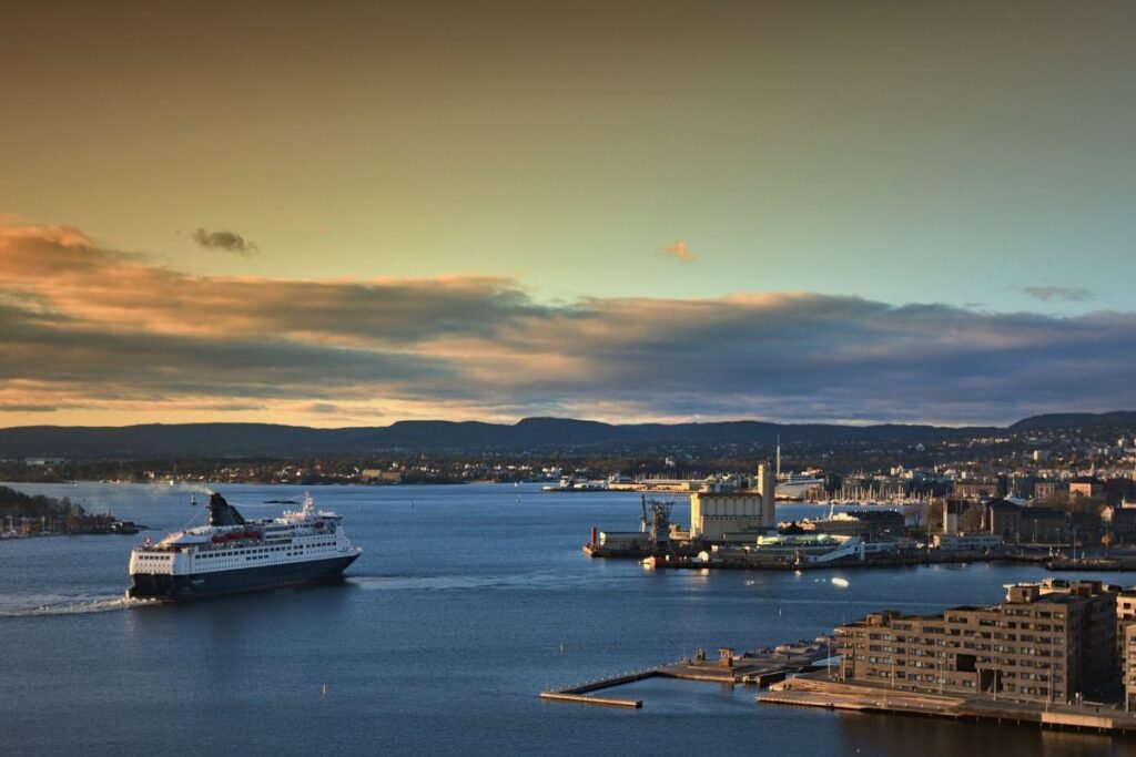 Eine Fähre in den früheren blau-weißen DFDS-Farben verlässt bei Sonnenuntergang den Hafen von Oslo, mit der Stadt und den Hügeln im Hintergrund