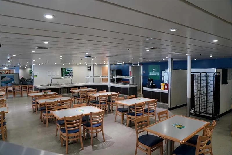 Selbstbedienungsrestaurant von Irish Ferries mit leeren Holzstühlen mit blauen Sitzen, weißen Tischen und einer Buffetlinie im Hintergrund