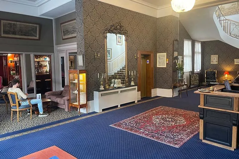 Lobby des Hotel Meurice in Calais mit blauem Teppich, einem vergoldeten Spiegel über dem Heizkörper und einer Bar hinter einer offenen Tür