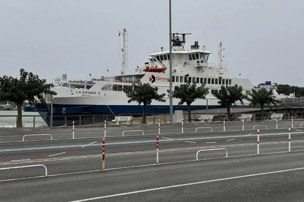 Die weiße Fähre La Gironde aus Bordeaux am Kai, gesehen über leere Wartespuren mit rot-weißen Pfosten und Ketten, unter grauem Himmel