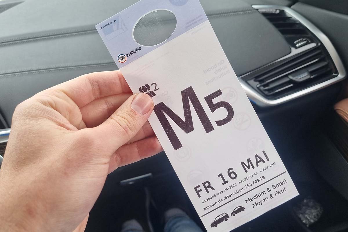 Hand mit einer Spiegelkarte von LeShuttle im Auto: ein großer Buchstabe mit Zahl, der Reisetag und unten rechts die Klasse Medium & Small mit zwei kleinen Autos