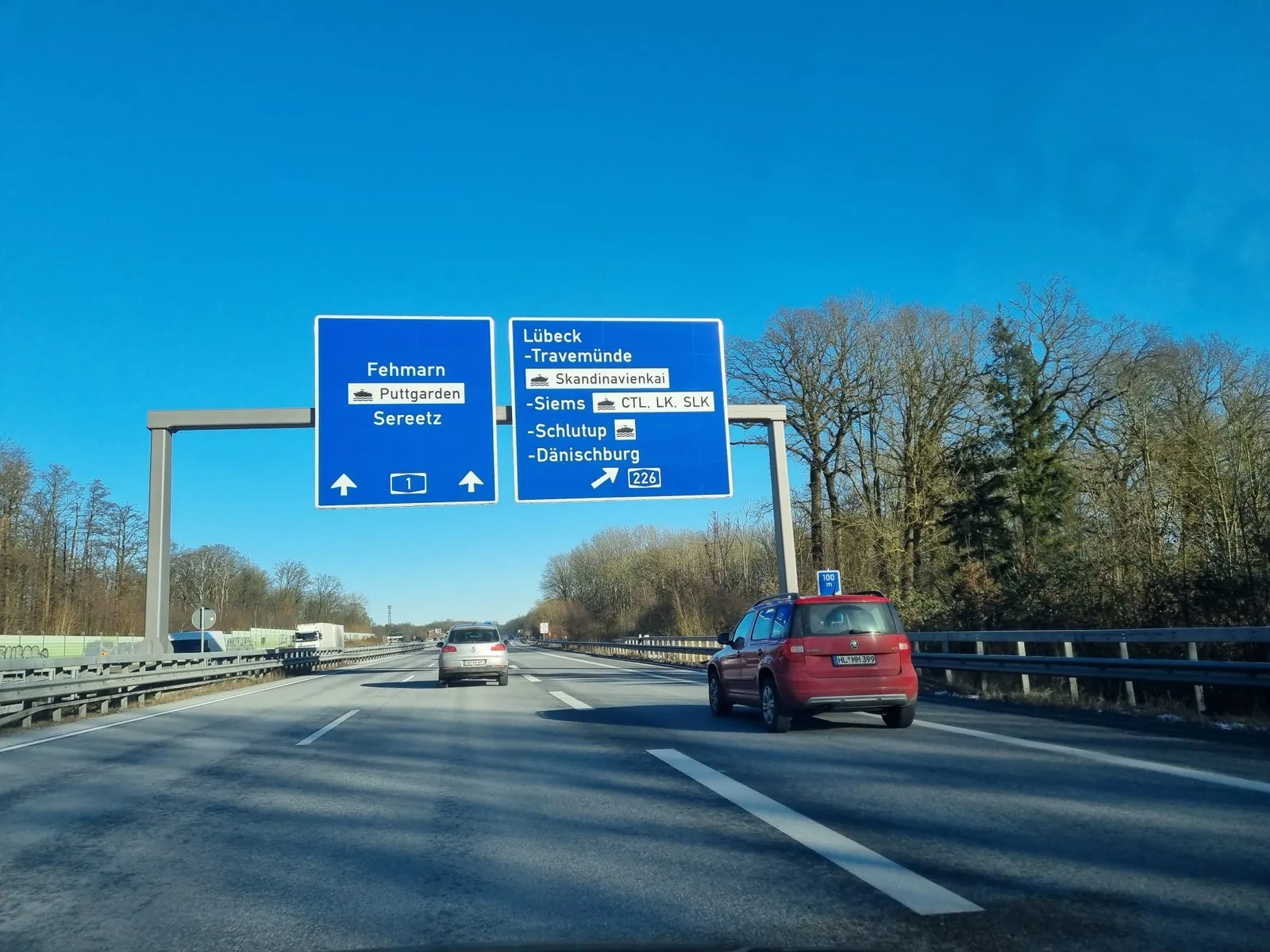 Blaues Schilderportal über einer deutschen Autobahn mit Fehmarn, Puttgarden mit Fährsymbol und Sereetz, darunter zwei Autos auf der Straße