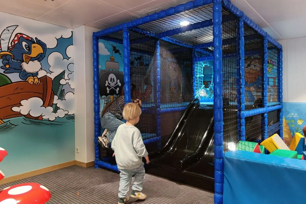 Ein Kind vor dem blauen Klettergerüst mit Rutsche im Pirates Club, mit einer Piratenmalerei an der Wand