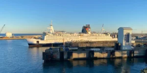Eine weiße Fähre von Scandlines am Kai in einem Hafen, im tiefen Winterlicht unter strahlend blauem Himmel, mit einer Kaimauer und einem grauen Aufzugsschacht im Vordergrund