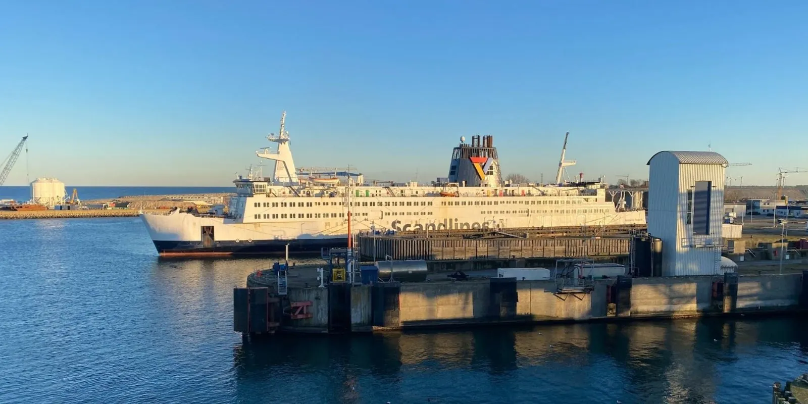 Eine weiße Fähre von Scandlines am Kai in einem Hafen, im tiefen Winterlicht unter strahlend blauem Himmel, mit einer Kaimauer und einem grauen Aufzugsschacht im Vordergrund