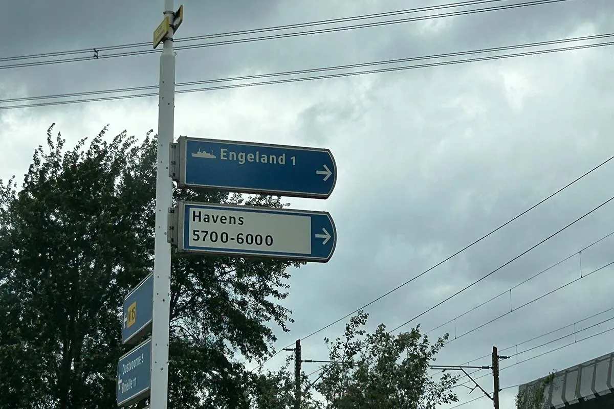 Ein blaues Wegweiserschild an der Straße mit einem Schiffchen und dem Wort Engeland, darunter ein weißes Schild mit Havens 5700-6000, unter bewölktem Himmel mit Oberleitungen