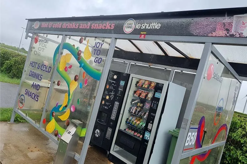 Glashäuschen auf dem Aufstellplatz mit einem Snackautomaten und einem Kaffeeautomaten, darüber der Text hot and cold drinks and snacks