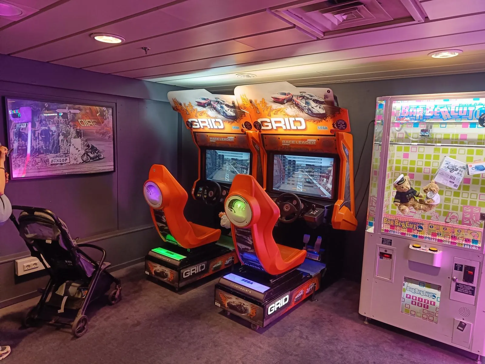 Zwei orangefarbene Rennspielautomaten und ein Greifautomat in der Spielhalle an Bord, mit einem Kinderwagen daneben