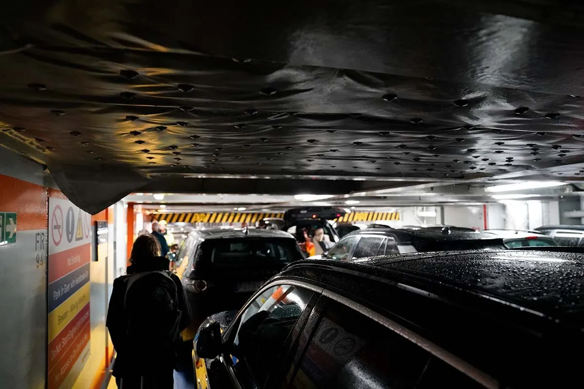 Autos dicht an dicht auf einem Autodeck mit einem tief hängenden Zwischendeck darüber; Passagiere gehen mit Taschen zwischen den Autos hindurch