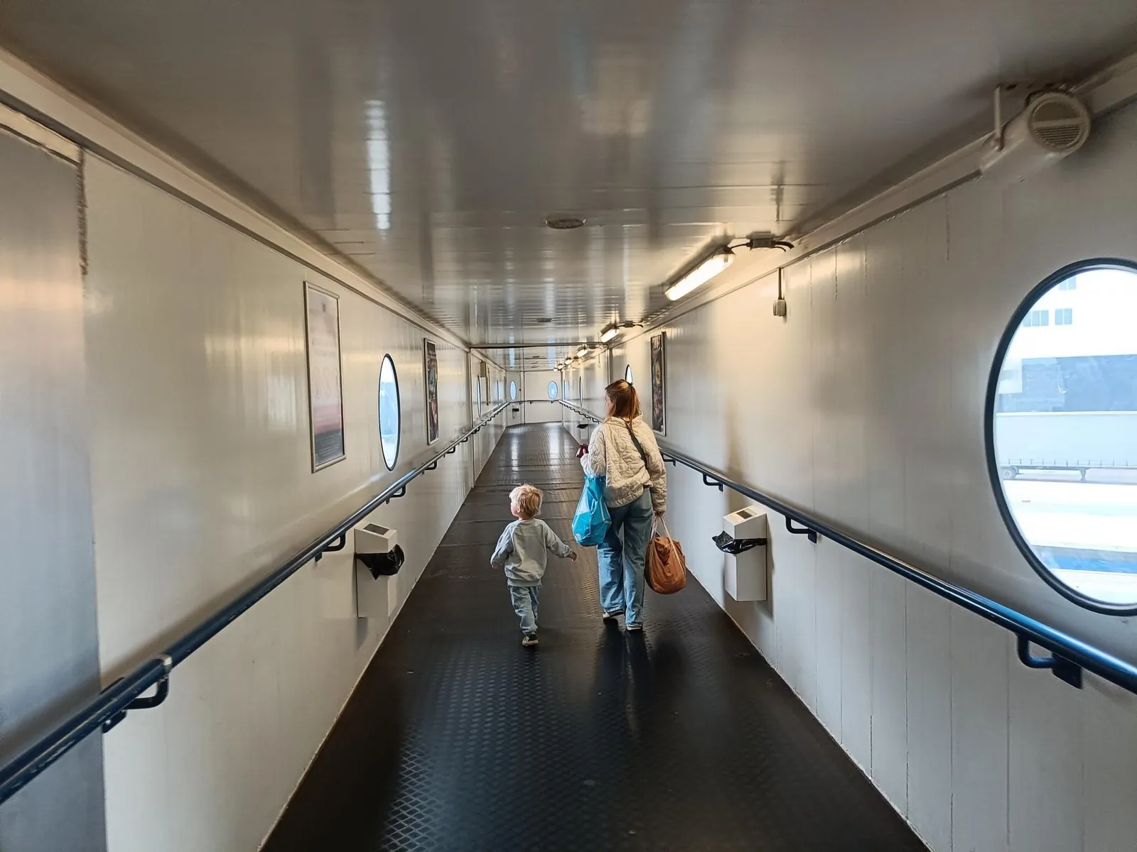 Ein Erwachsener mit Taschen und ein Kleinkind gehen durch die überdachte Gangway vom Terminal zum Schiff