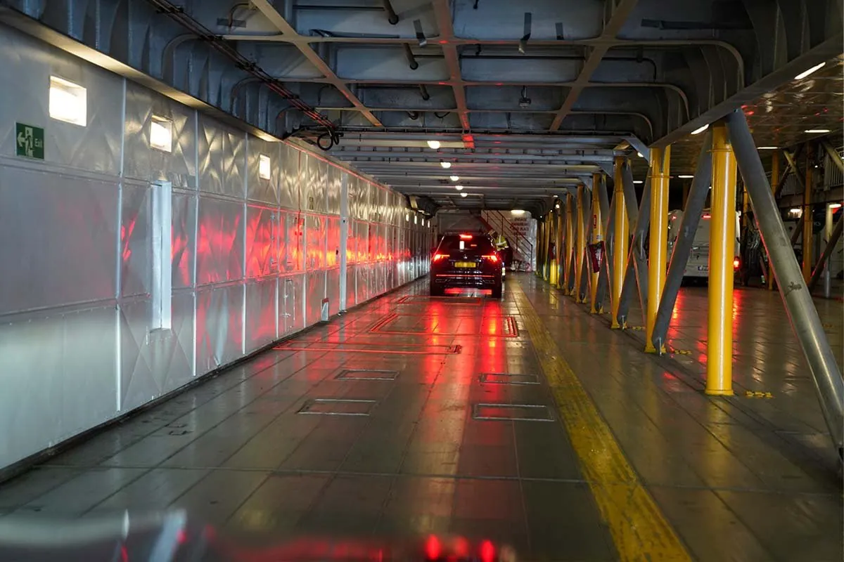 Autodeck an Bord mit silbernen Wandpaneelen, gelben Stützpfeilern und einem Auto mit leuchtenden Bremslichtern, die sich auf dem Boden spiegeln