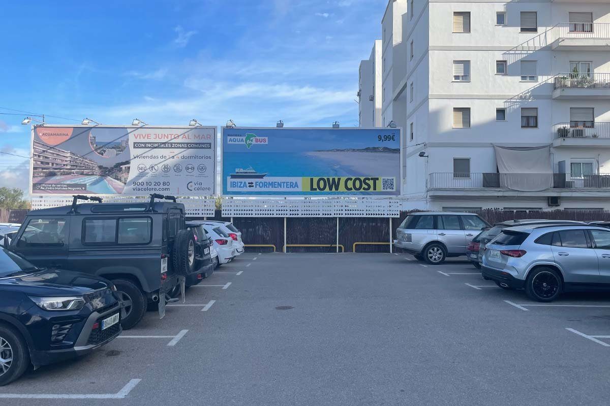 Parkplatz mit Autos, dahinter zwei Werbetafeln; die rechte wirbt für die Fähre nach Formentera mit einem Betrag von 9,99 Euro