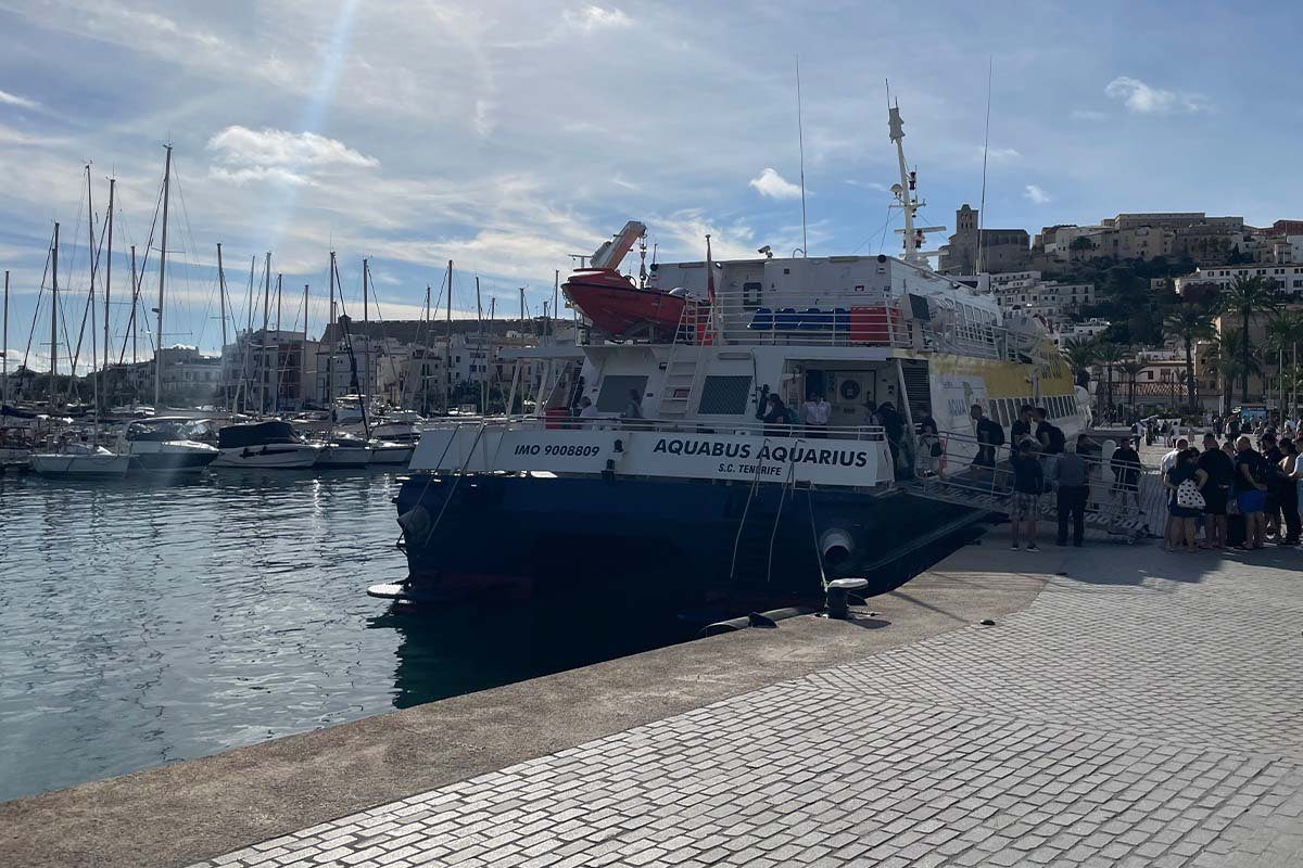 Die Schnellfähre Aquabus Aquarius mit dunkelblauem Rumpf am Kai im Hafen von Ibiza, daneben eine Schlange wartender Passagiere, auf dem Hügel dahinter die Altstadt Dalt Vila