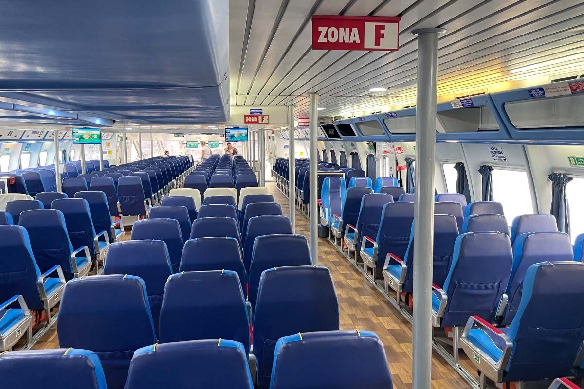 Passagierdeck einer Schnellfähre mit langen Reihen blauer Sitze, einem Boden in Holzoptik und einem roten Schild mit Zona F an der Decke