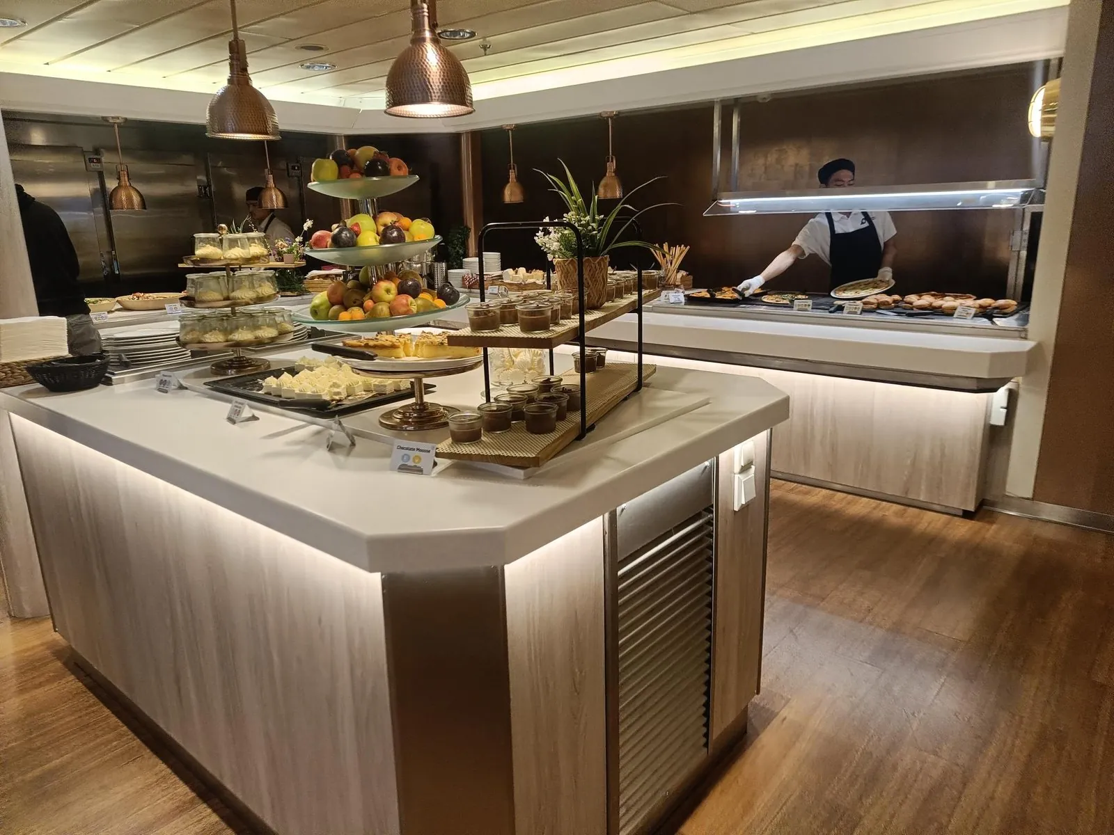 Die Buffetinsel mit Desserts und Obst im Buffetrestaurant, dahinter ein Koch an der warmen Theke