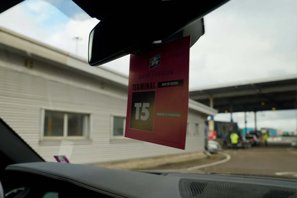 Rote Karte von Irish Ferries mit der Aufschrift Terminal T5 am Innenspiegel eines Autos; durch die Windschutzscheibe die Check-in-Häuschen und ein Mitarbeiter in gelber Weste