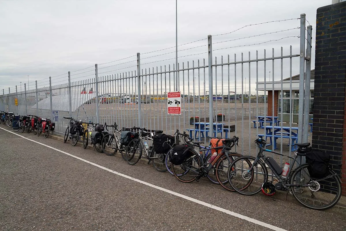Eine Reihe vollgepackter Fahrräder mit Packtaschen an einem Stahlzaun beim Terminal von Harwich, dahinter Picknicktische