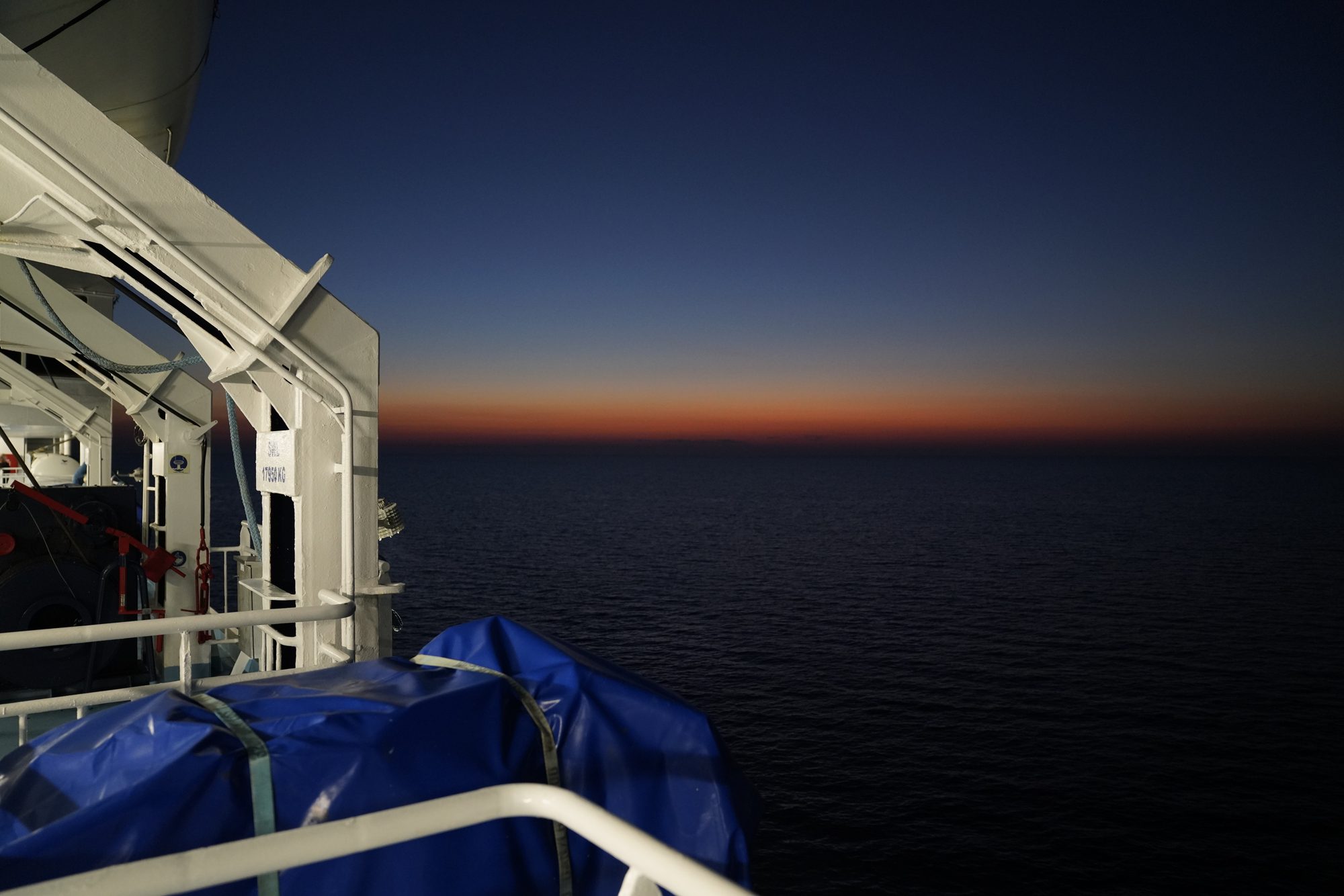 Blick vom Deck über das dunkle Meer mit einem orangen Streifen am Horizont nach Sonnenuntergang, links ein Rettungsboot