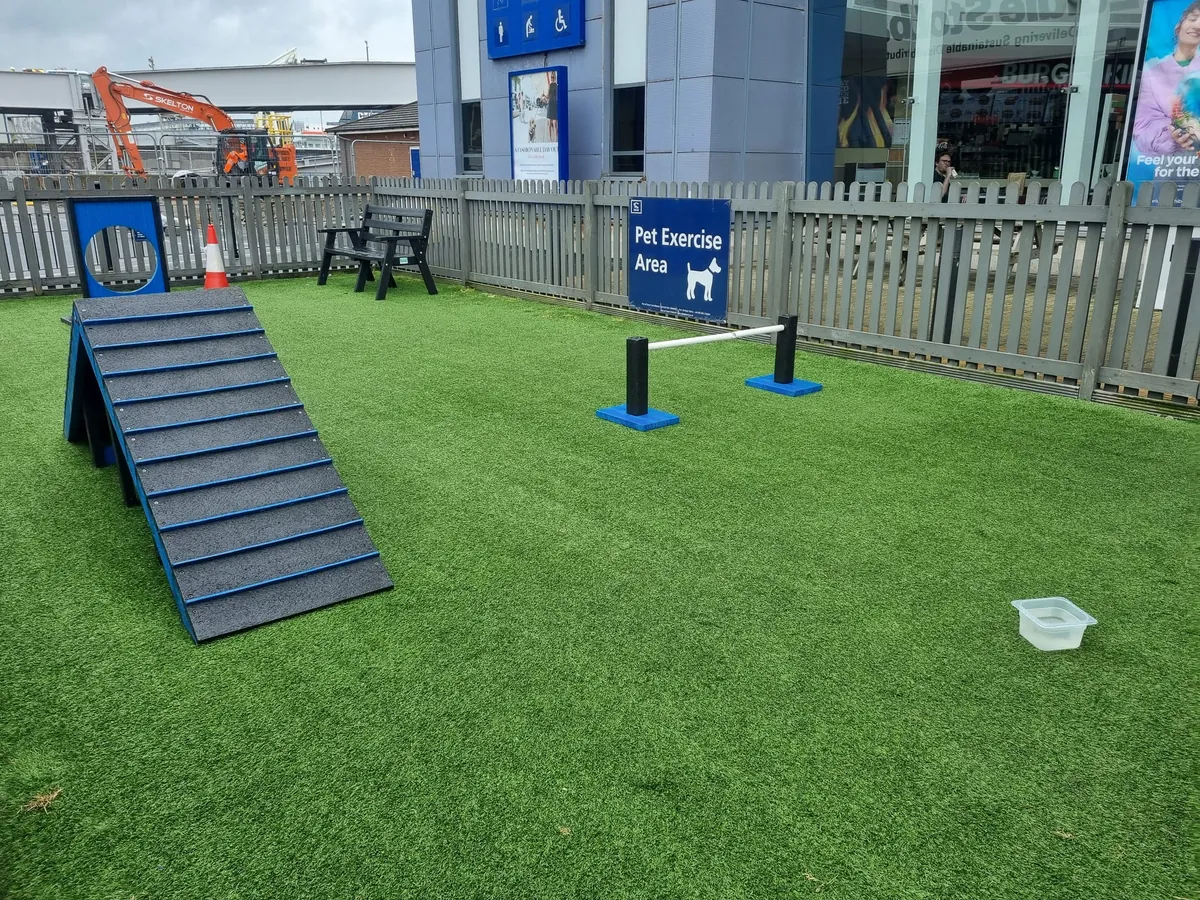 Eine Auslauffläche mit Kunstrasen hinter einem grauen Holzzaun, mit einer Rampe, einer Sprunglatte, einer Tafel mit einem runden Loch, einer Bank, einem Wassernapf und dem Schild Pet Exercise Area, vor einem Terminalgebäude
