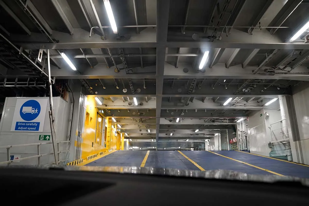 Das leere Autodeck der Fähre, durch die Windschutzscheibe gesehen, mit gelben Fahrlinien und einem Schild Drive carefully, adjust speed