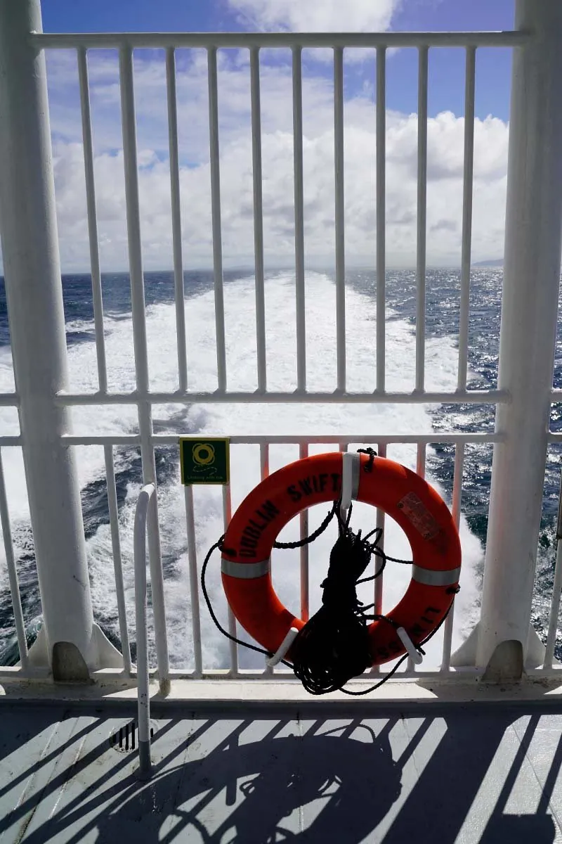 Ein oranger Rettungsring mit der Aufschrift Dublin Swift an einem hohen weißen Zaun auf dem Achterdeck, dahinter das schäumende Kielwasser