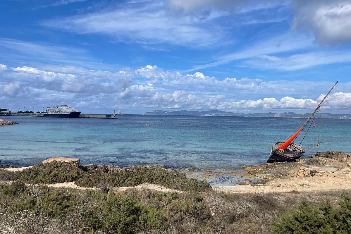 Blick von der felsigen Küste Formenteras über klares türkisfarbenes Wasser auf eine Fähre von Trasmapi an der Hafenmole von La Savina, am Horizont die Küste von Ibiza