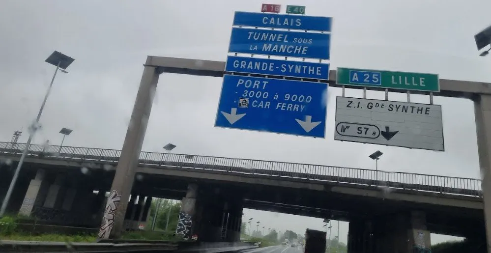 Schilderbrücke über der A16 mit blauen Schildern nach Calais, Tunnel sous la Manche und Port Car Ferry, durch eine nasse Windschutzscheibe gesehen