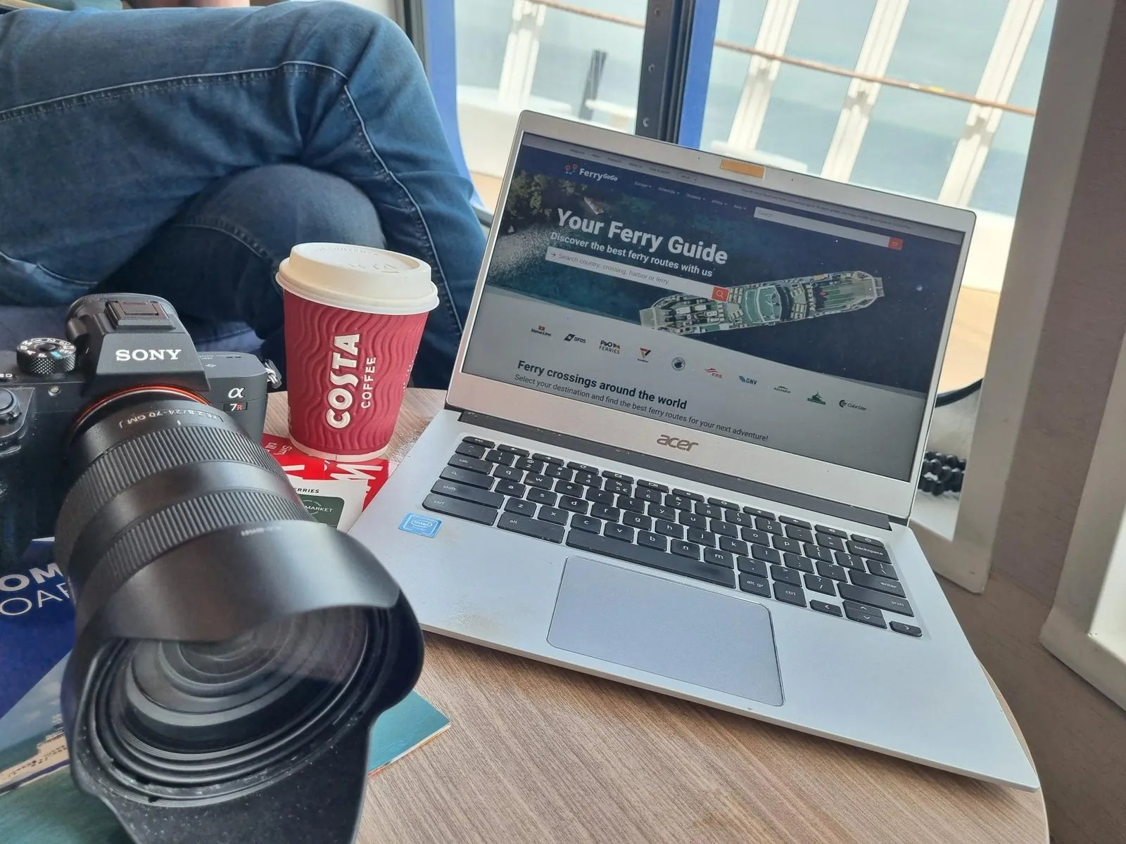 Laptop mit der Website von Ferrygogo auf einem Tisch am Fenster, daneben ein Becher Costa-Kaffee und eine Kamera mit Teleobjektiv
