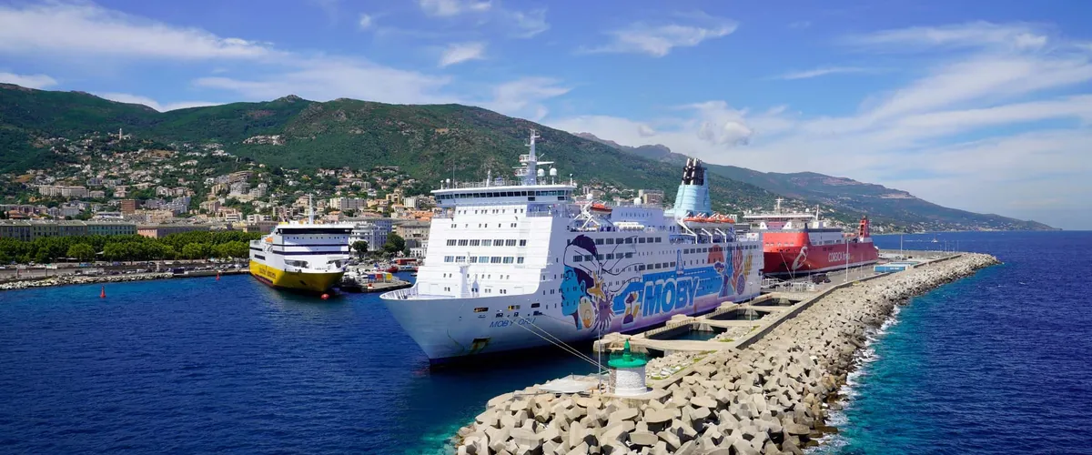 Der Hafen von Bastia vom Meer aus, mit drei Fähren am Kai: eine gelbe von Corsica Sardinia Ferries, die weiße Moby Orli mit der Looney-Tunes-Bemalung und eine rote von Corsica Linea, vorn der Wellenbrecher und dahinter die Berge von Cap Corse