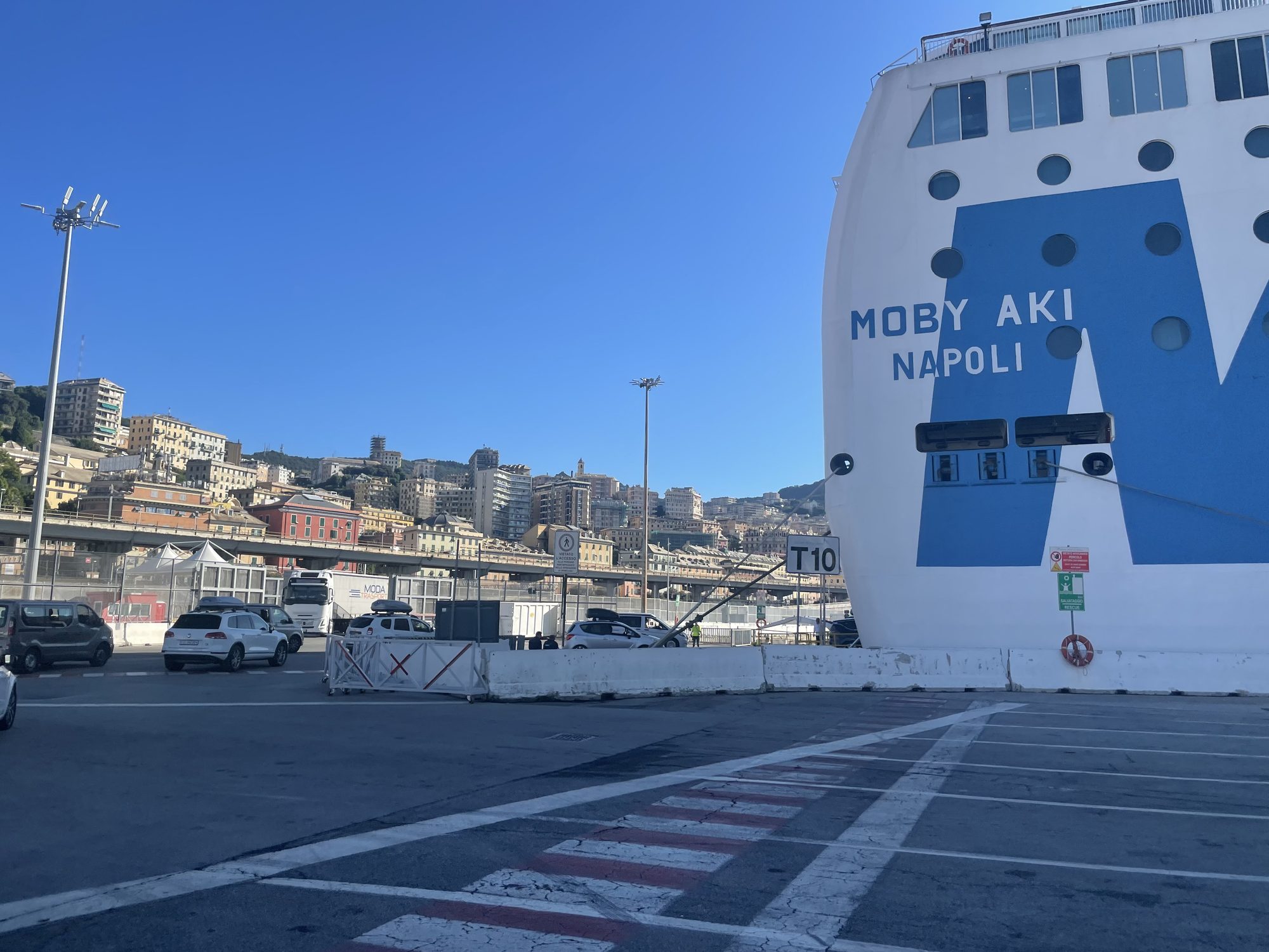 Der Bug der Fähre Moby Aki mit dem Heimathafen Napoli am Kai T10 in Genua, dahinter die Stadt auf den Hügeln