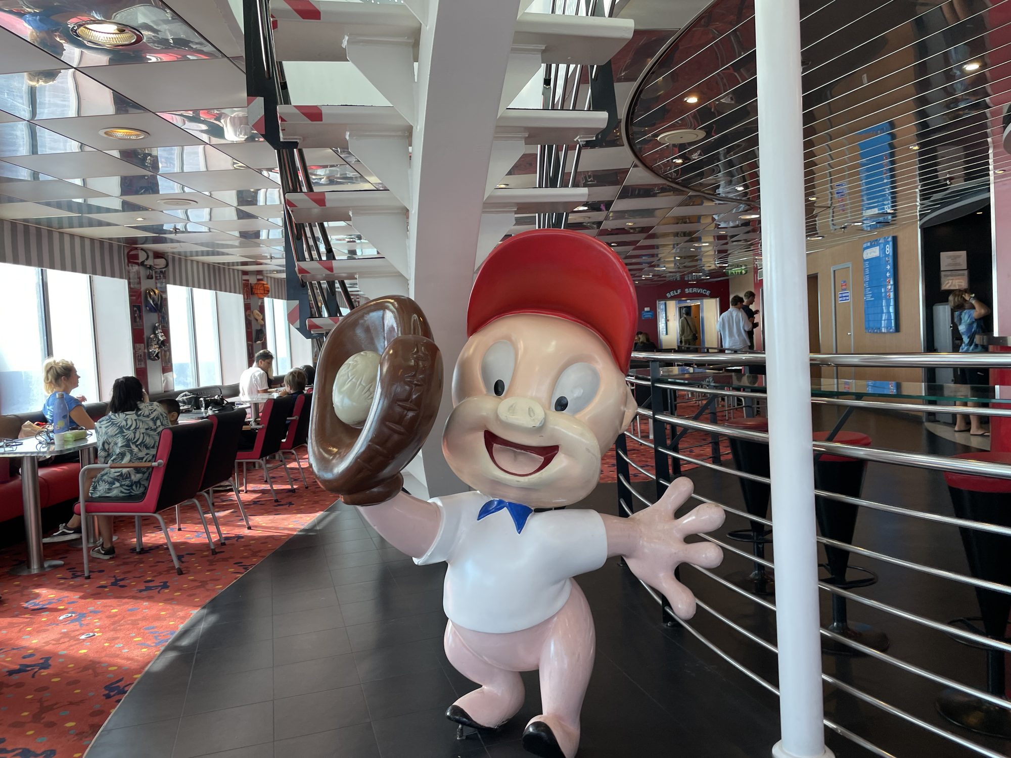 Eine lebensgroße Figur von Porky Pig mit Mütze und Baseballhandschuh neben dem Eingang der Selbstbedienung an Bord