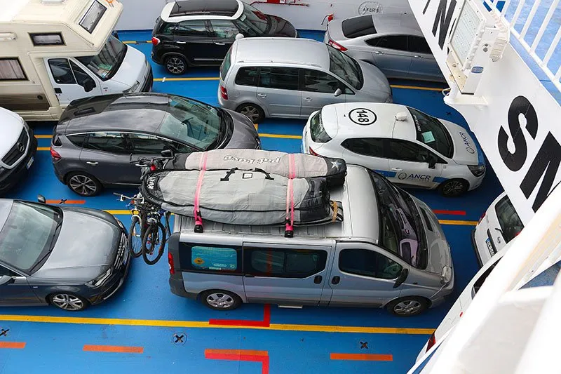 Ein volles Autodeck von oben: Autos dicht an dicht zwischen gelben Linien, ein weißes Wohnmobil in der Ecke und ein Bus mit Surfbrettern auf dem Dach und Fahrrädern auf dem Träger