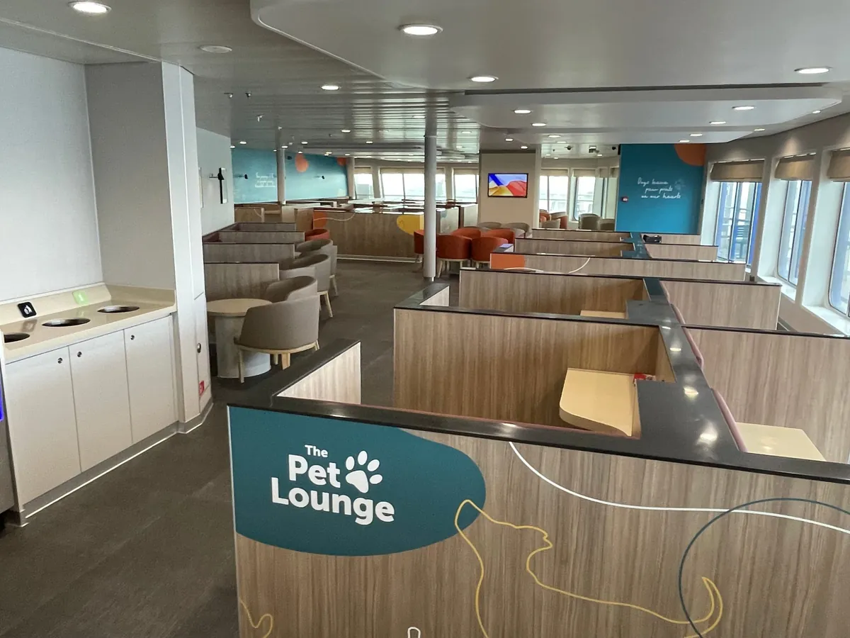 The Pet Lounge an Bord: Sitzgruppen mit Holztrennwänden dazwischen, Stühle und runde Tische, petrolblaue Wände mit dem Schriftzug Dogs leave paw prints on our hearts und Fenster an der Seite