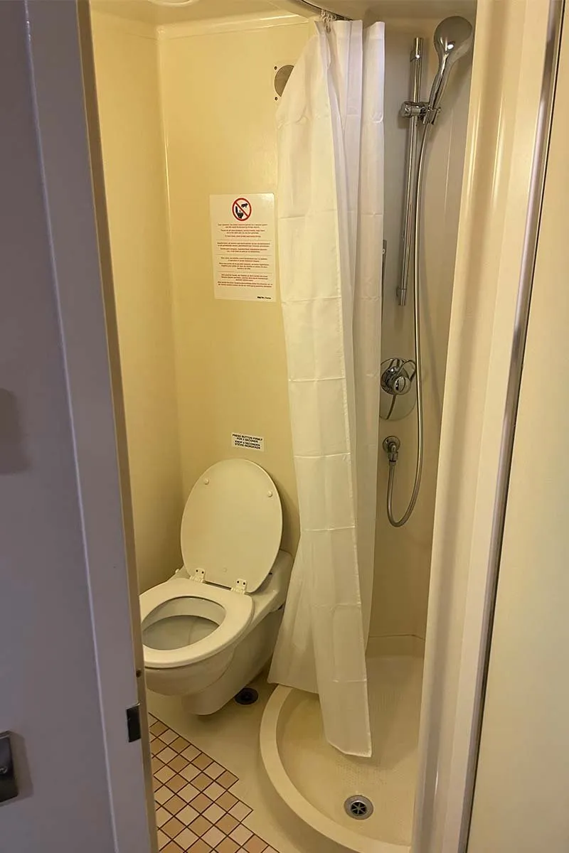 Das kleine Bad der Kabine mit einem Hänge-WC, einer runden Duschwanne mit weißem Duschvorhang und einer Handbrause, alles aus cremefarbenem Kunststoff