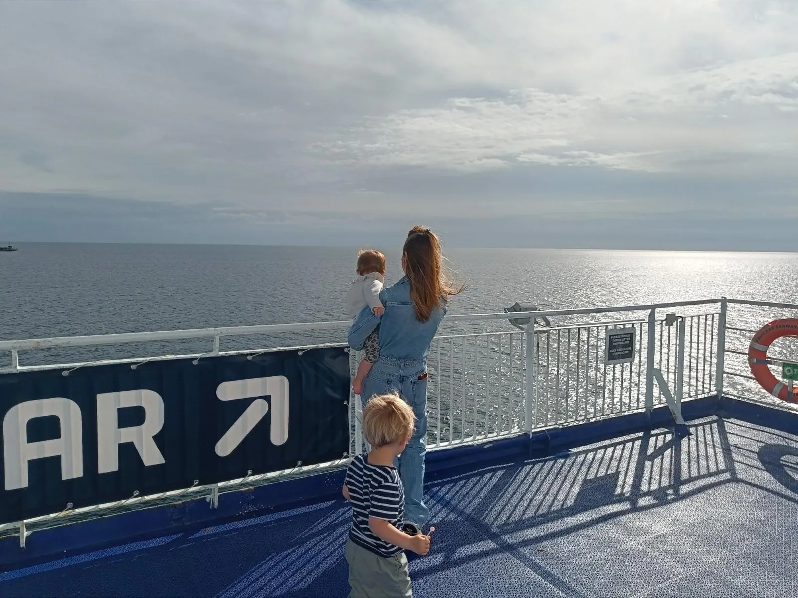 Ein Erwachsener und zwei Kinder schauen vom Außendeck über das Meer, neben einem Schild zur Sky Bar