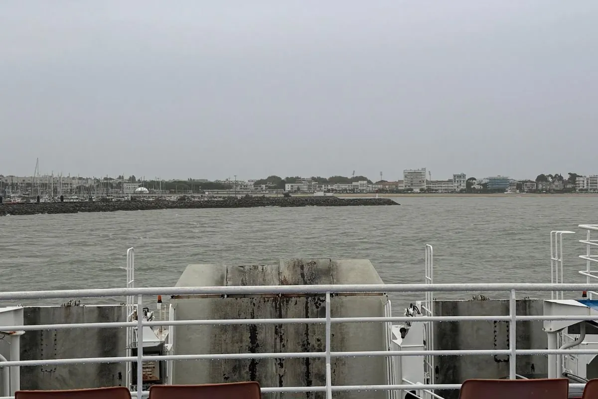 Blick vom Deck über die Reling und die Rettungsinseln auf graues Wasser, mit einer steinernen Mole und den Wohnblocks und dem Jachthafen eines Badeorts auf der anderen Seite