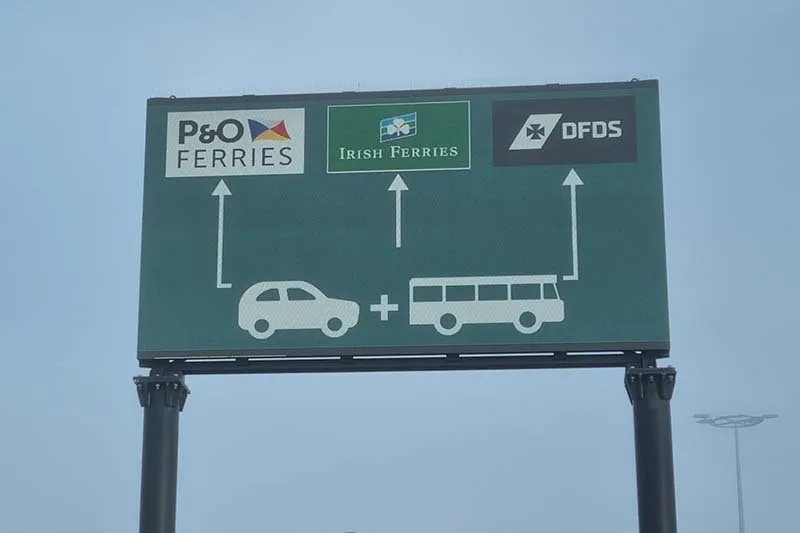 Grünes Verkehrsschild über der Straße zum Hafen von Calais mit den Logos von P&O Ferries, Irish Ferries und DFDS und Pfeilen für Autos und Busse